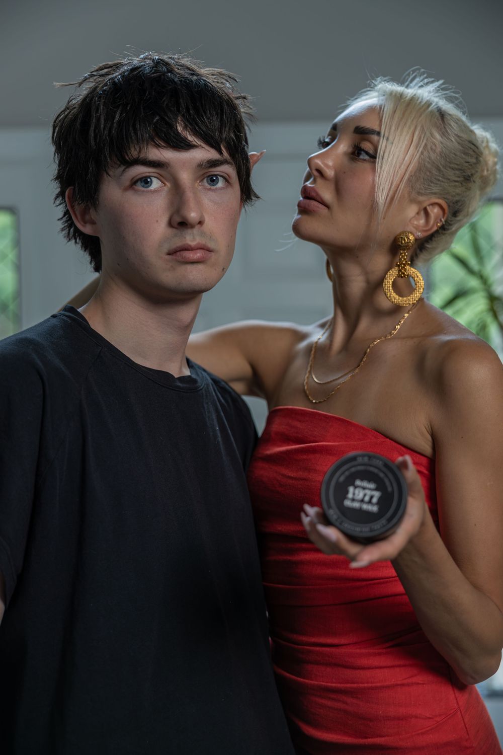 Young man in black tee and blonde woman in red dress holding hair pomade jar pose together indoors.