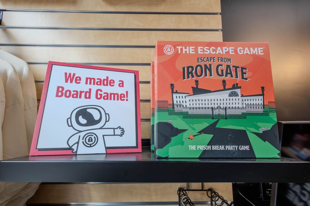 The Escape Game Iron Gate prison break party board game displayed on a shelf next to a We Made a Board Game sign.