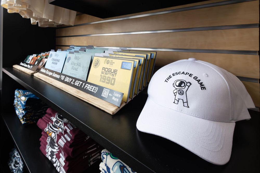 White The Escape Game branded trucker hat displayed on shelf beside folded merchandise and gift cards in retail store.