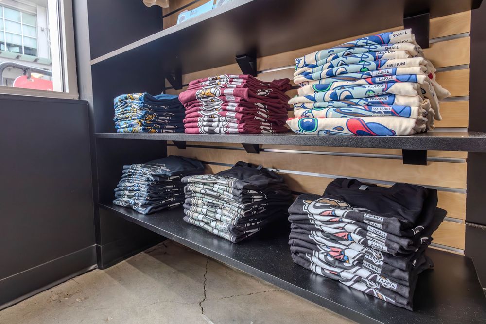 Folded t-shirts displayed on black retail shelves in a sports merchandise store with slatwall panels.