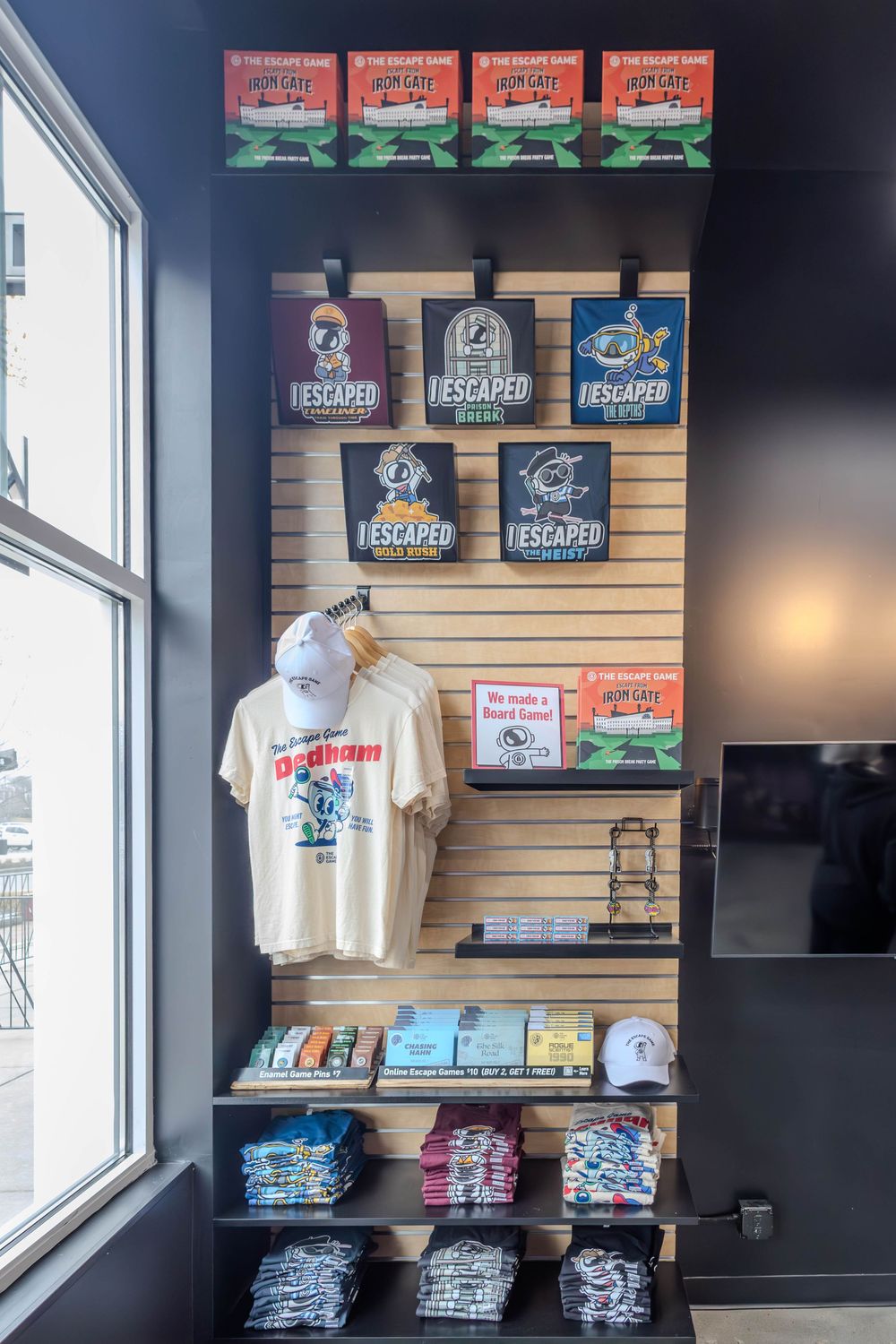 Escape Game merchandise display wall featuring t-shirts, hats, and "I Escaped" souvenir signs on dark slatwall shelving.