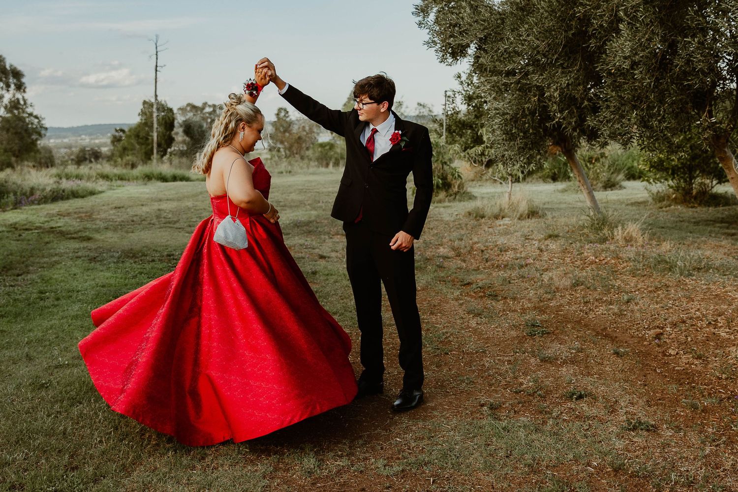 A beautiful Year 12 graduate in a gorgeous red gown twirling with her partner in a paddock for a professional formal photo session.