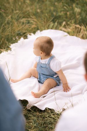 Baby in white clothing lying on a white blanket in grassy outdoor setting.