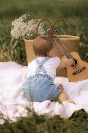 Toddler in denim overalls relaxing on white blanket next to acoustic guitar outdoors.