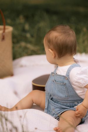 Baby wearing denim overalls resting on white blanket in natural outdoor setting.