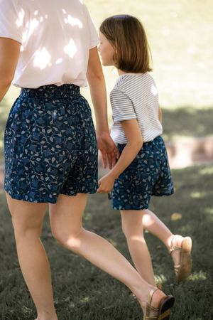 Mother and child wearing matching blue floral shorts walking in dappled sunlight.