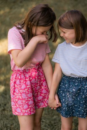 Young children wearing striped shirts and floral shorts hold hands while standing together outdoors.