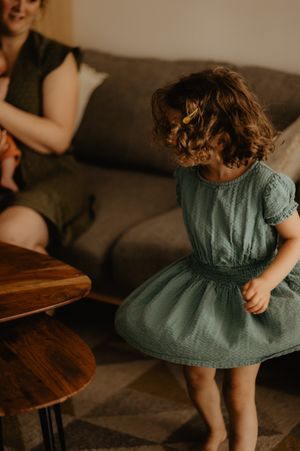 A child in a teal dress plays near a wooden table in warm indoor lighting.