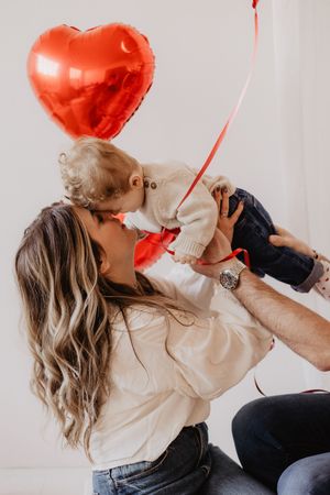 Romantic couple sharing kisses behind red heart-shaped balloons against white background.