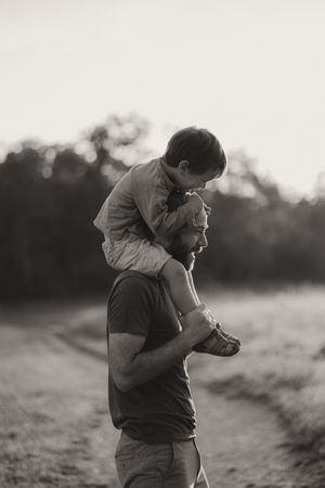 Black and white portrait of parent carrying child on shoulders outdoors.