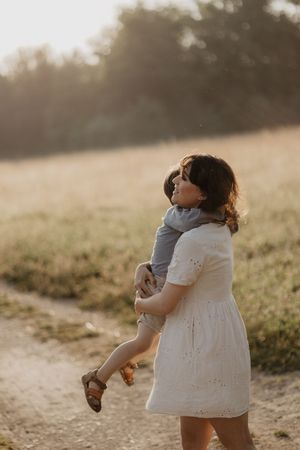 Person in white dress holding child while standing in sunlit meadow.