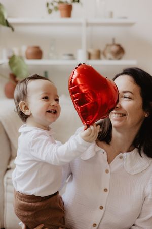 Parent and child sharing playful moments with red heart balloon in bright minimalist setting.