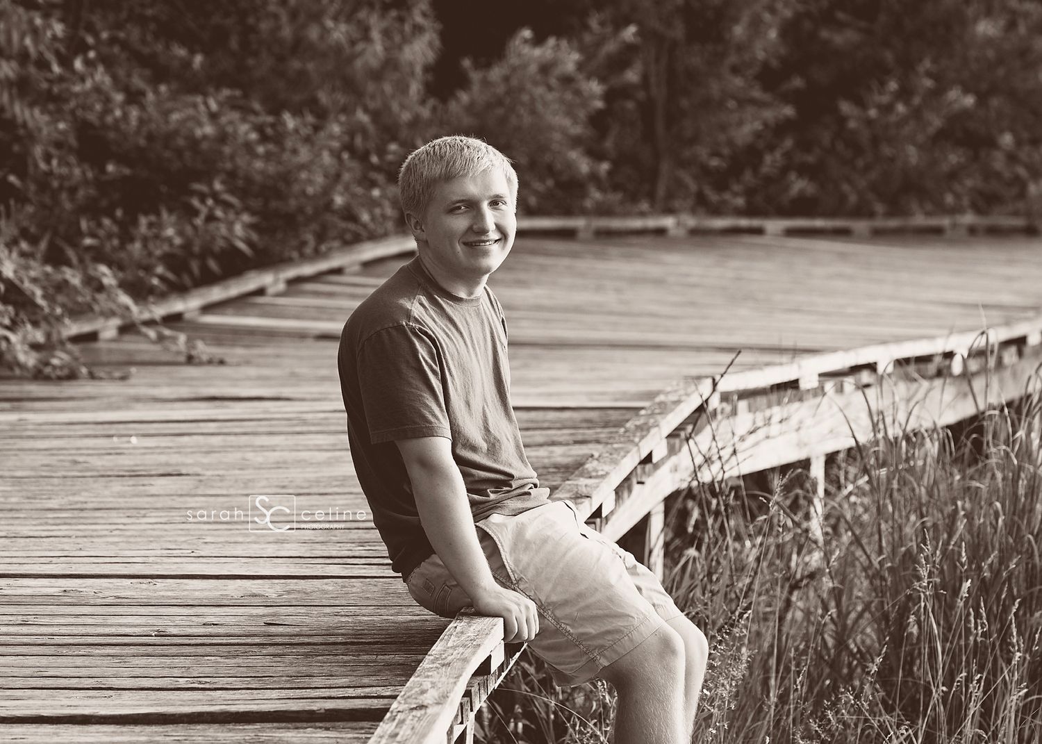 A child sits on a wooden dock wearing casual clothes while swinging their legs and looking down at the water below.