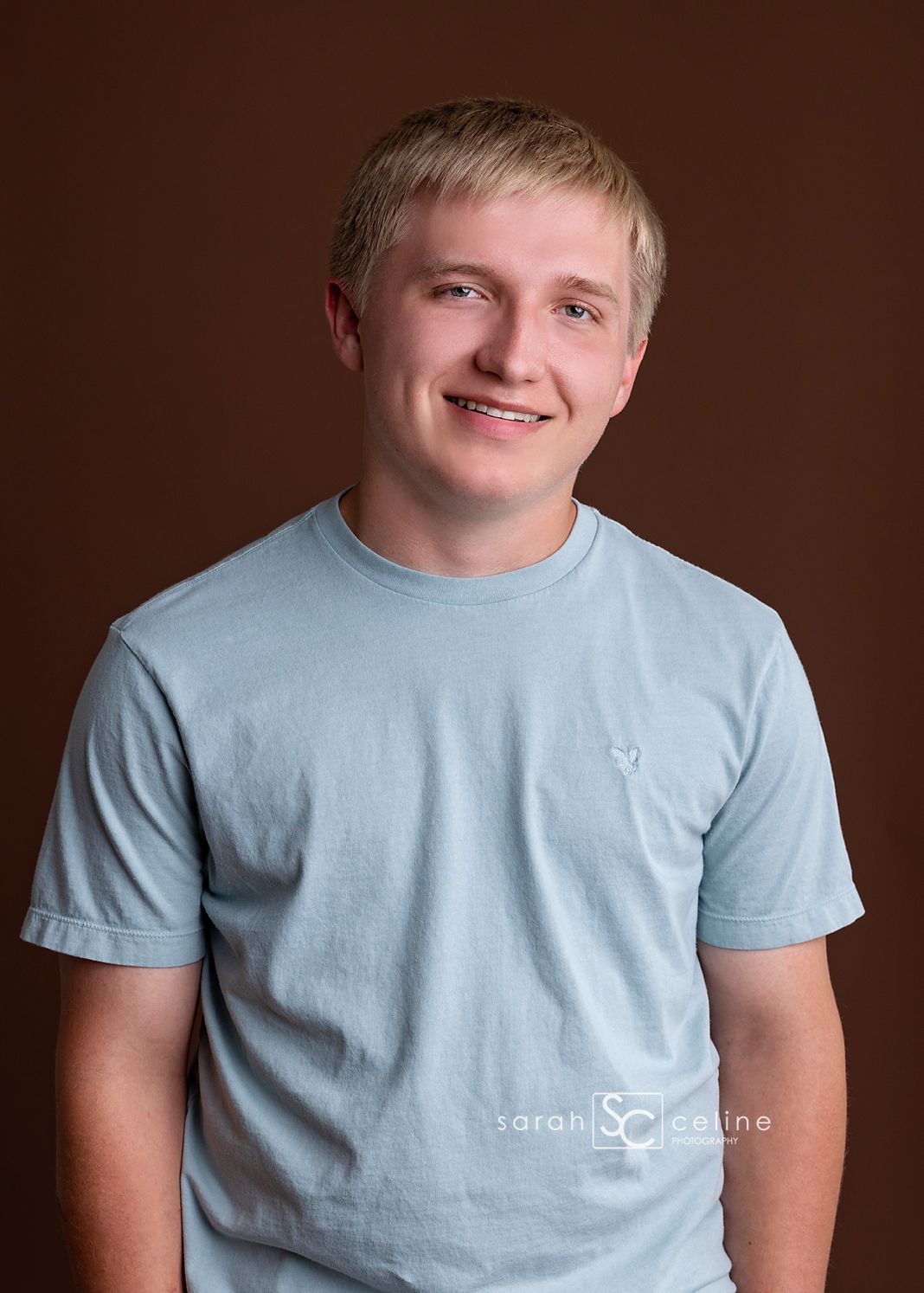 A young man wearing a light blue t-shirt is posing against a brown background during a photography session.