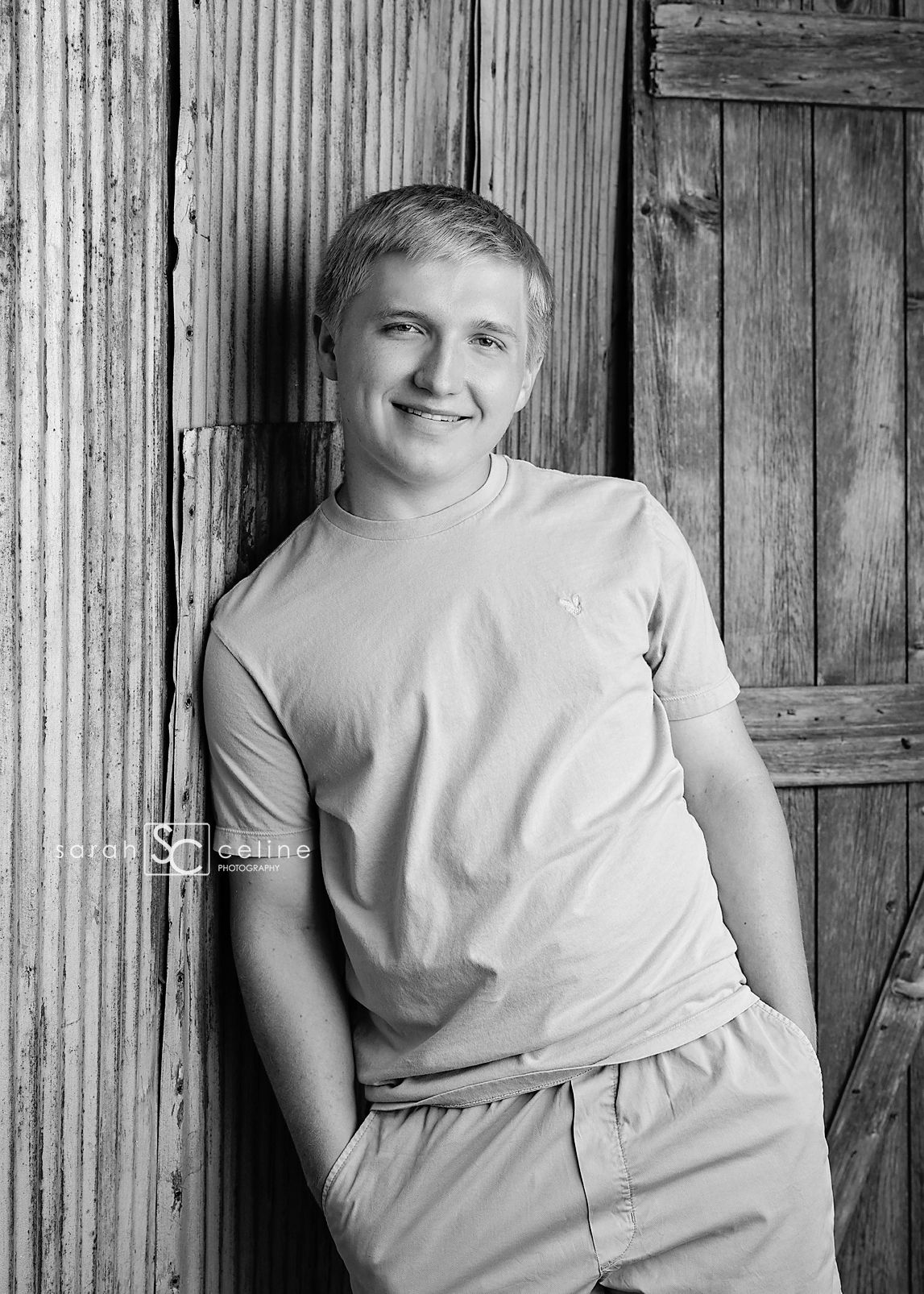 Black and white portrait of a young person in a light colored t-shirt standing against a rustic wooden wall.