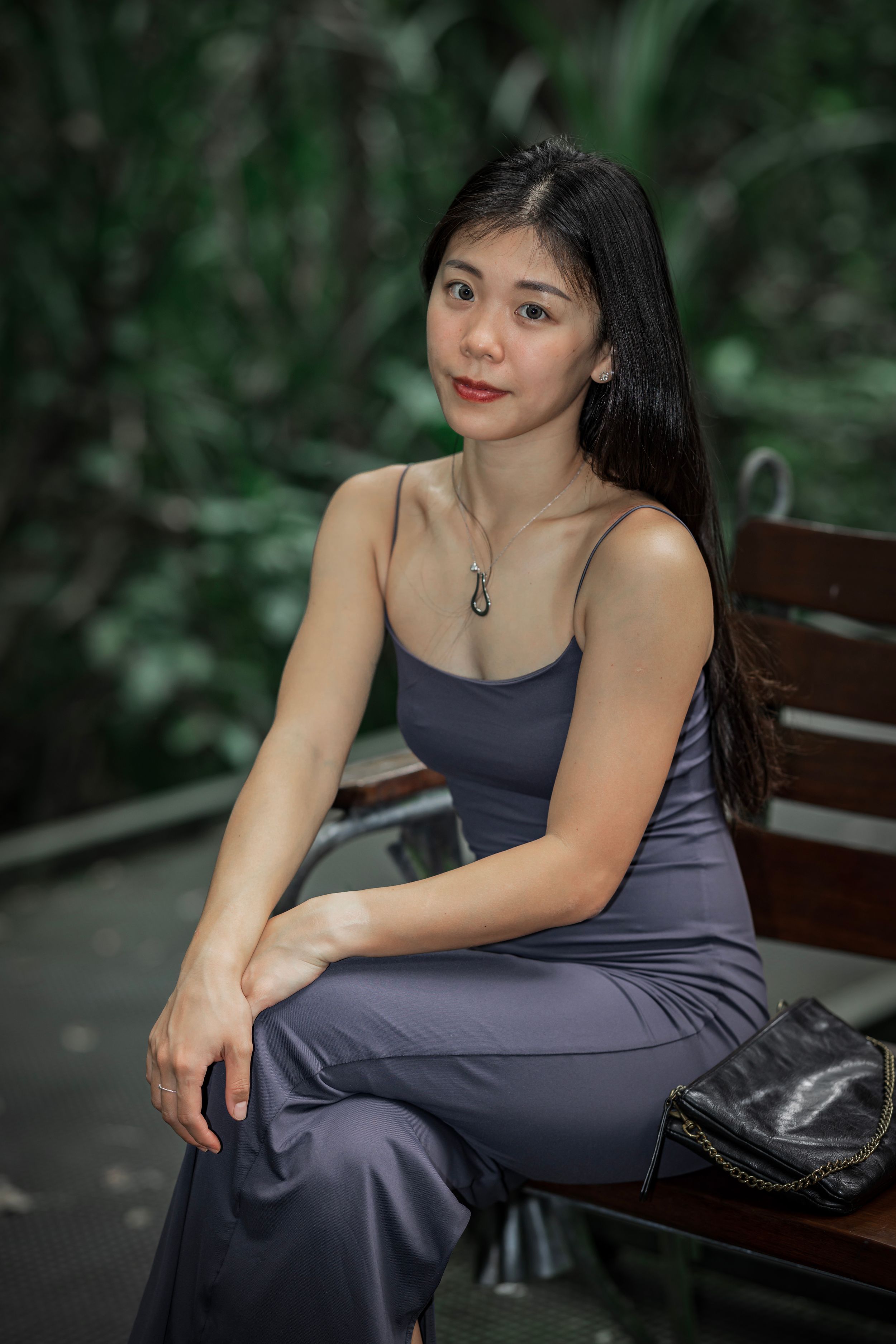 A person in a navy blue dress poses elegantly on a wooden park bench against a lush green natural background.