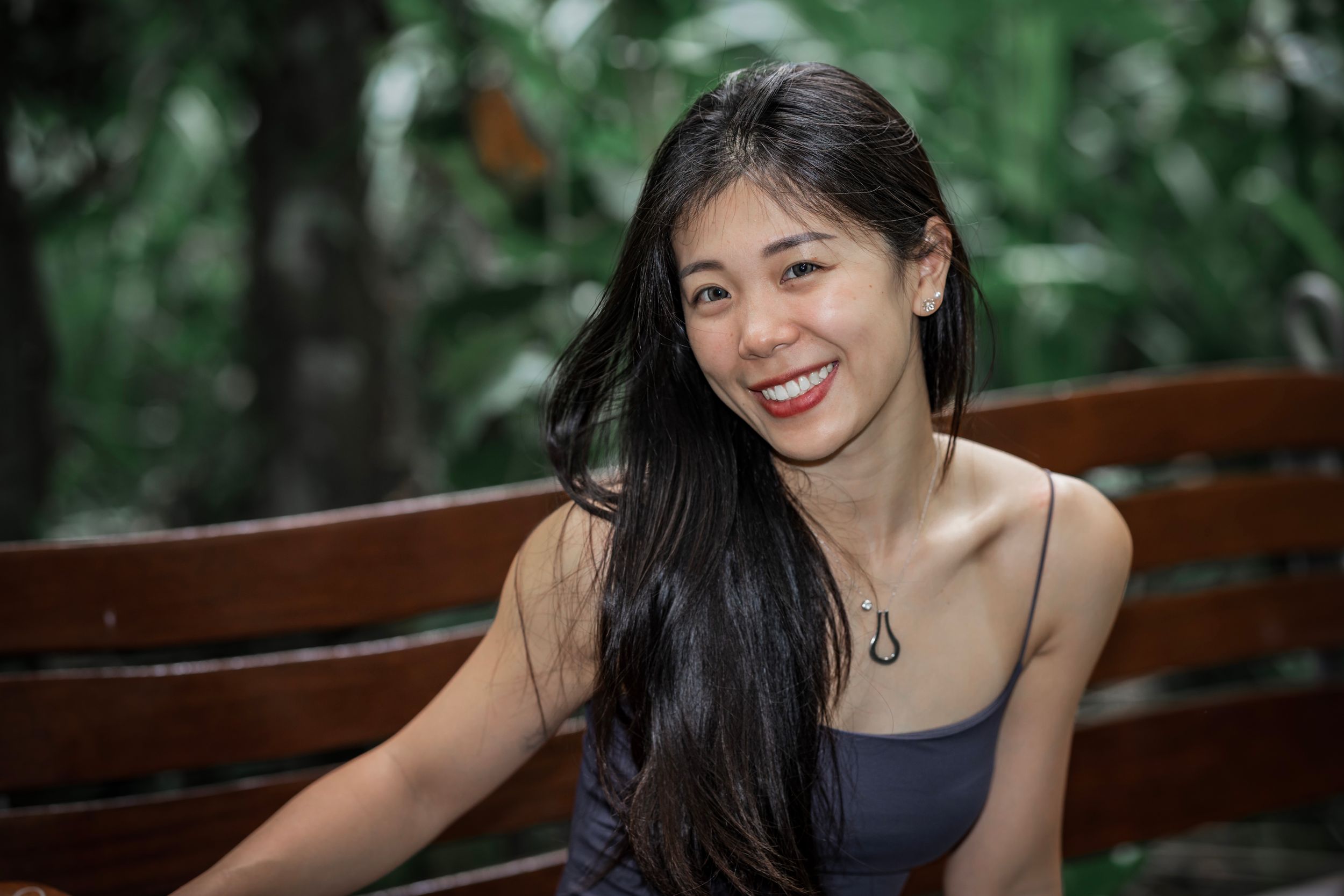 Smiling woman with long dark hair sitting on wooden bench outdoors with blurred green foliage background.