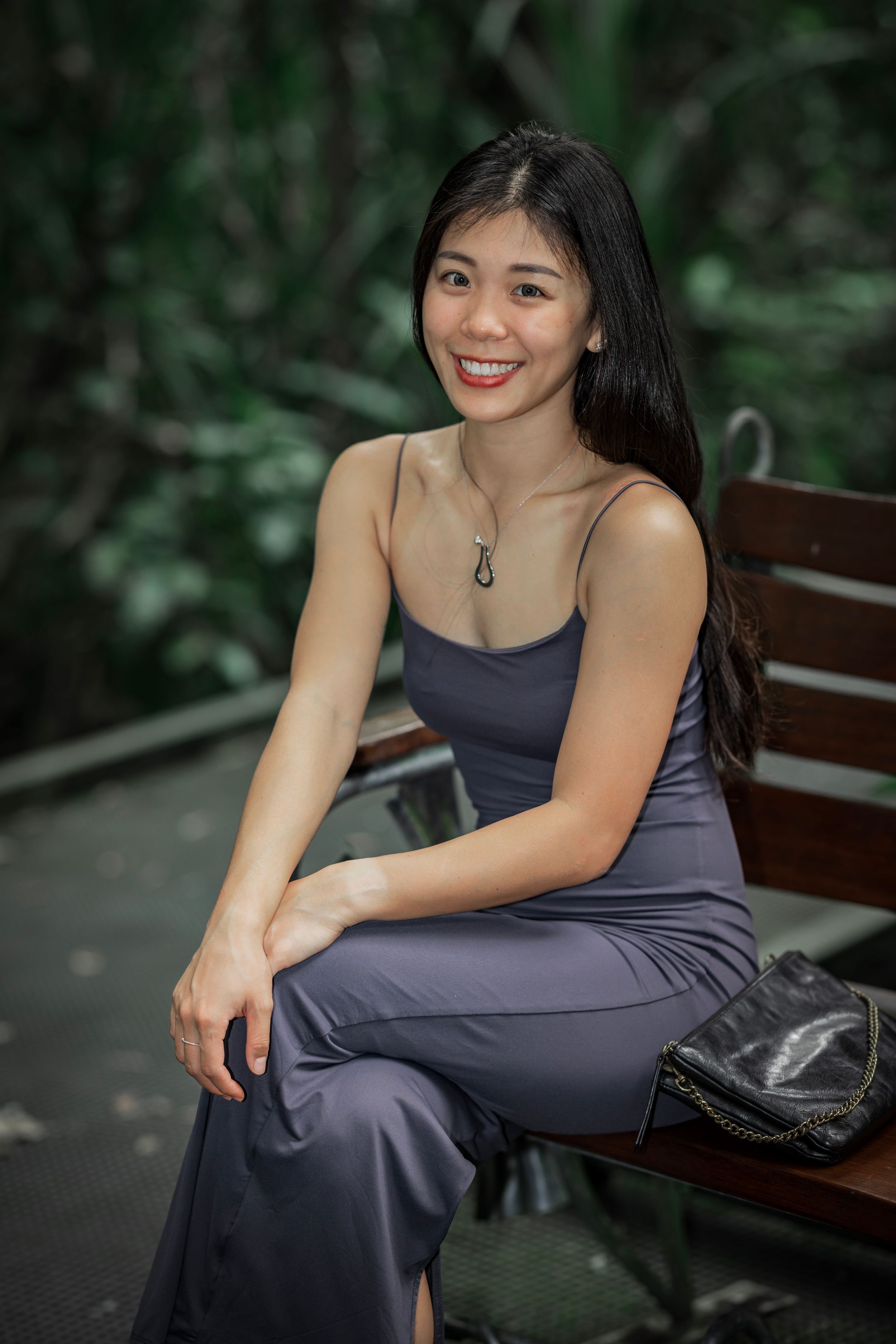 A person in a gray dress sits on a wooden bench outdoors against a lush green background.