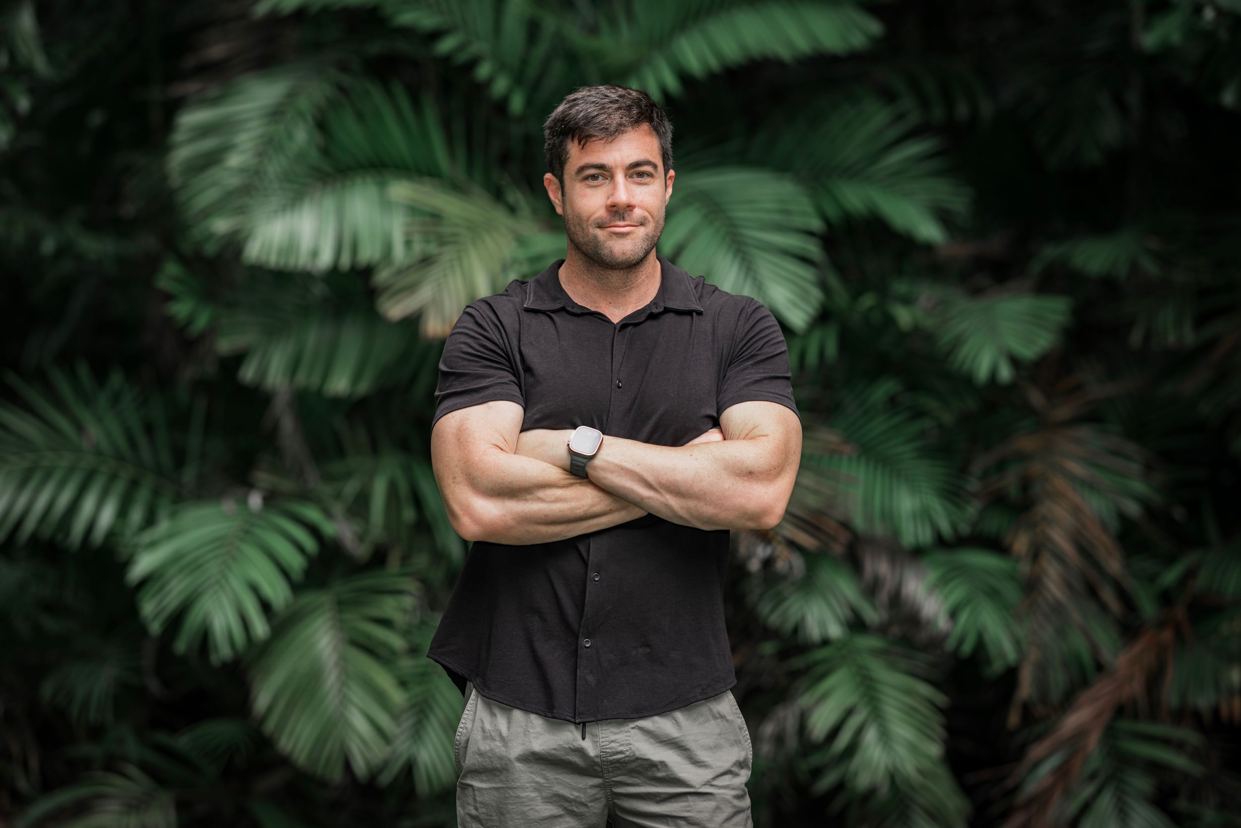 A figure in a black t-shirt stands outdoors against lush tropical greenery and palm fronds in a garden setting.
