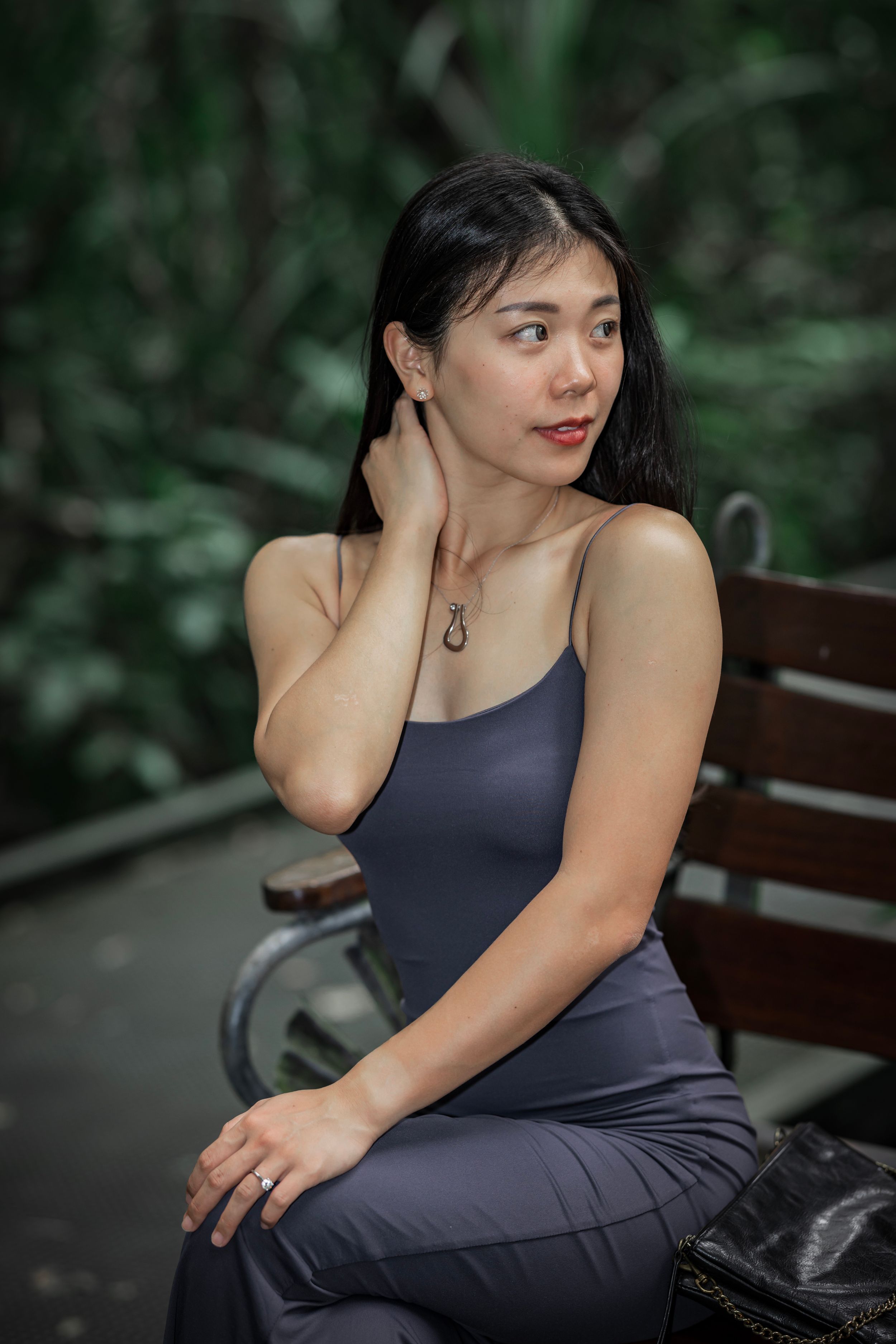 Woman in navy dress poses thoughtfully on wooden bench in garden setting with lush green foliage backdrop.