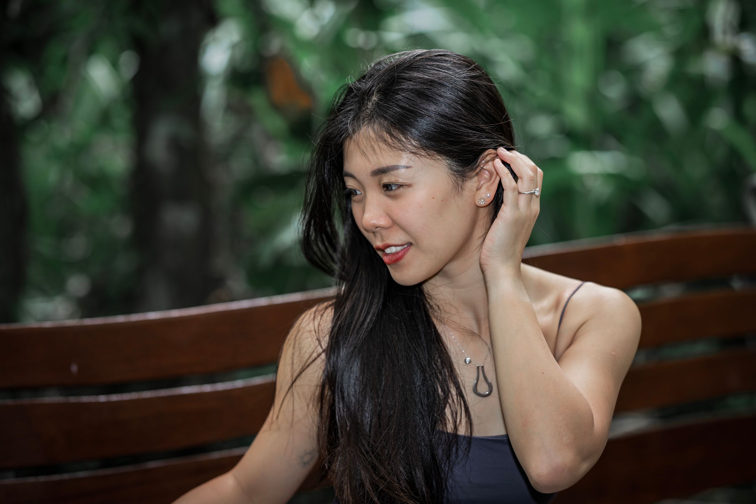 Women with long dark hair sit outside at a wooden table in a garden setting, smiling and looking down.