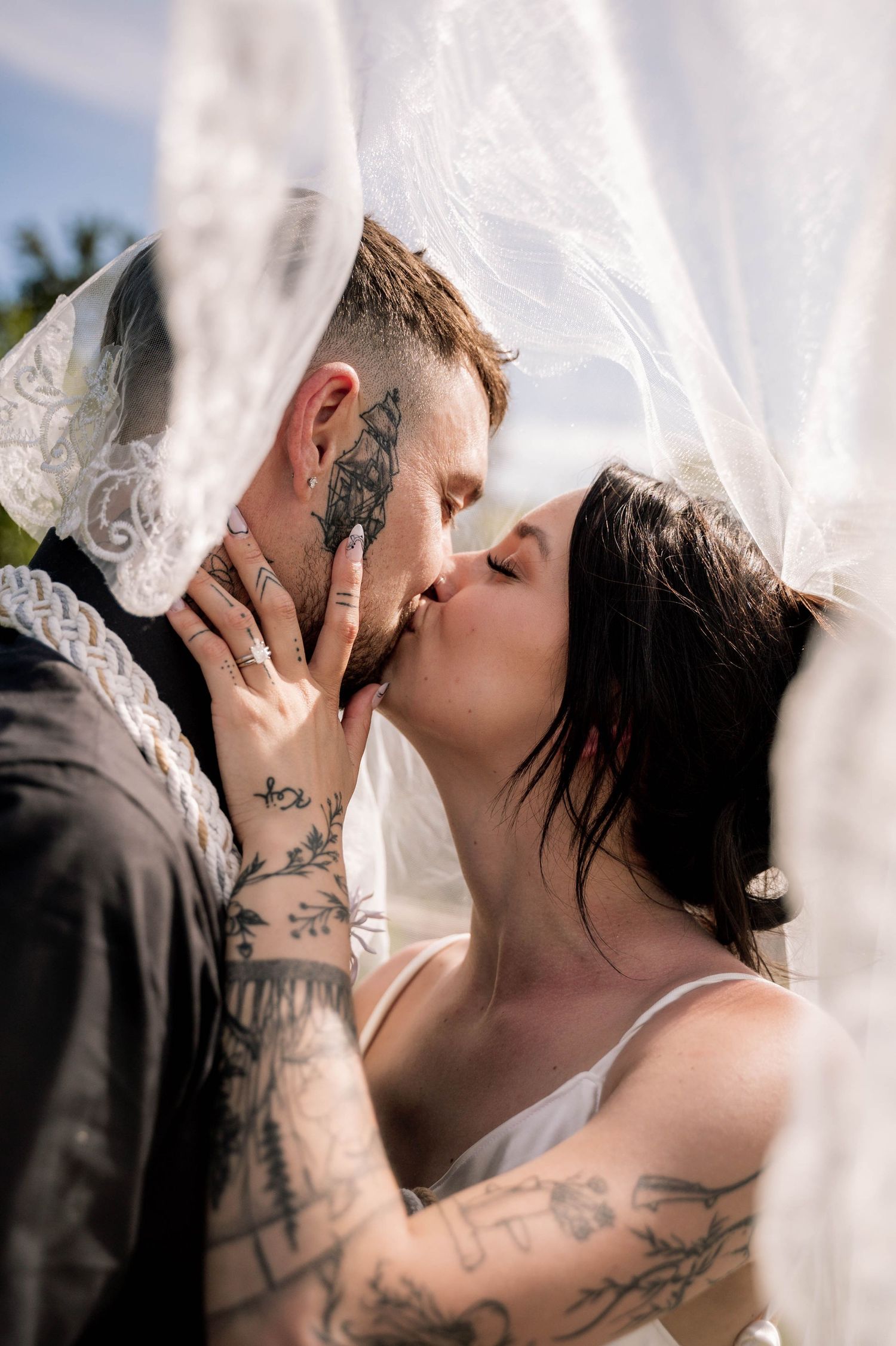 Romantic couple kissing under flowing white veil with visible tattoos in outdoor wedding photography.