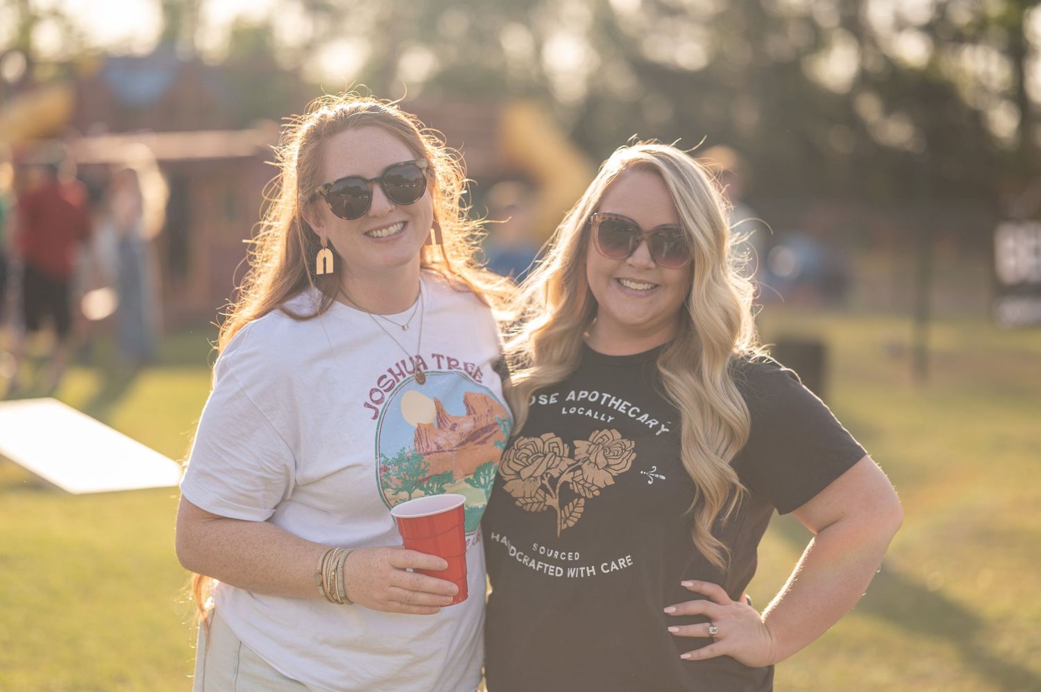Two friends wearing sunglasses and t-shirts pose together on a sunny day in a grassy field at golden hour.