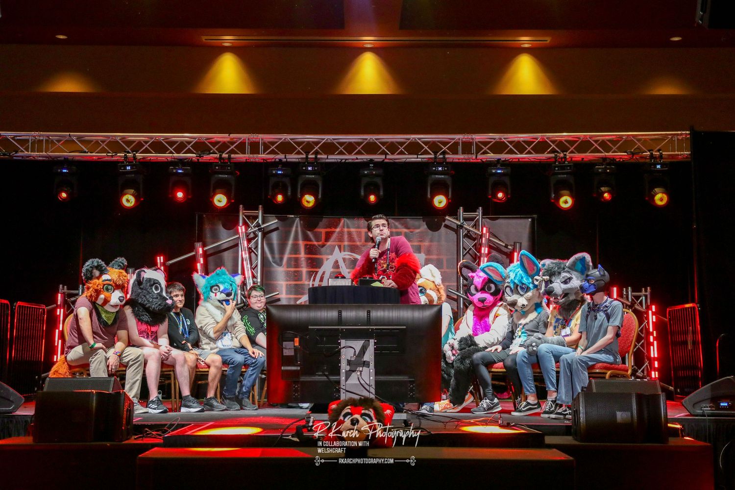Group of people experiencing a virtual reality attraction with headsets while seated in red chairs on stage under spotlights.