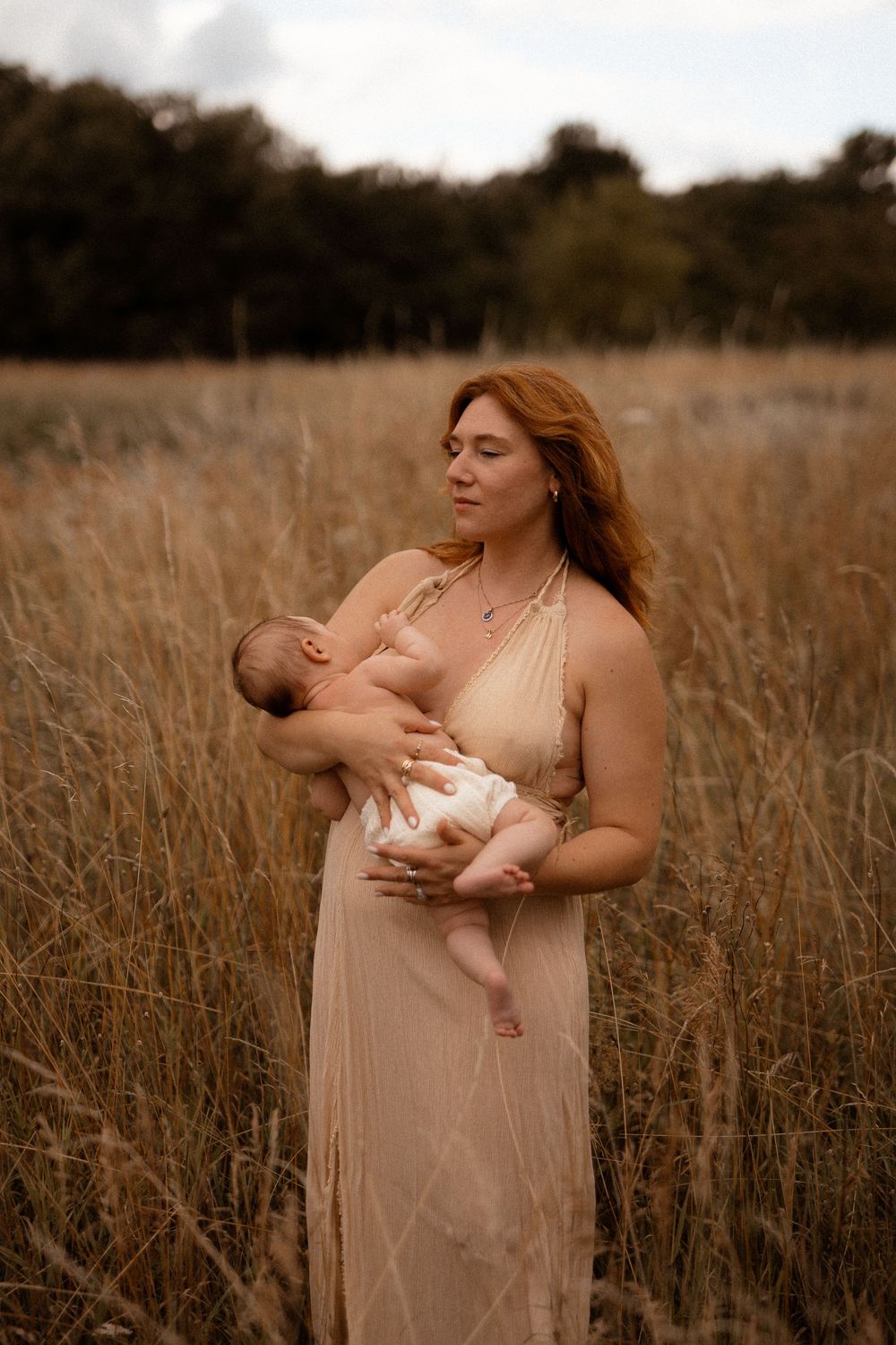 A tender moment captured in a golden field as a mother nurses her infant while wearing a flowing beige dress at sunset.