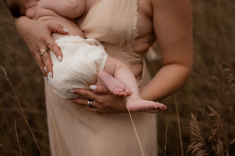 Tiny newborn baby feet rest gently in parent's hands against soft beige dress in outdoor nature setting.