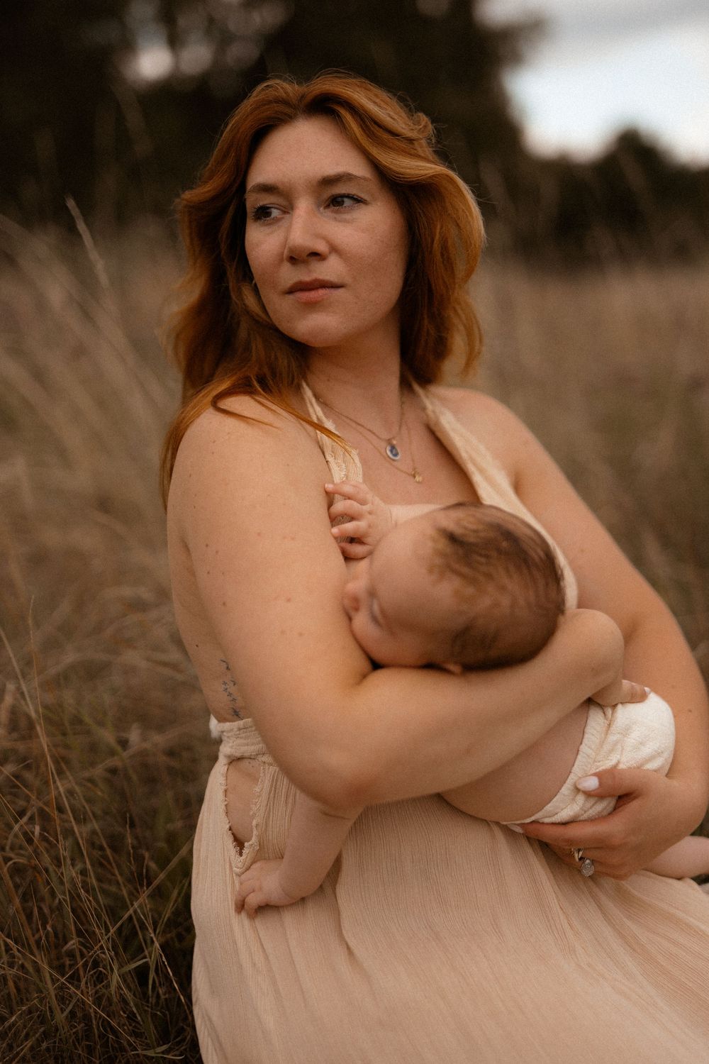 A mother in a pink dress nurses her baby in a natural outdoor setting at sunset.