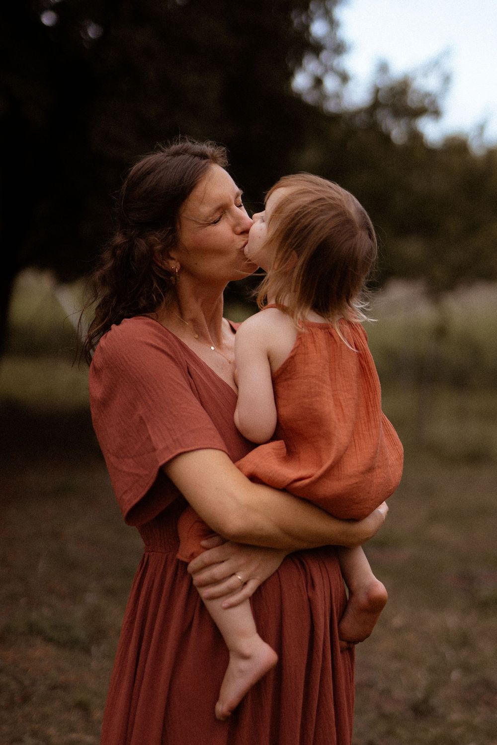 A tender moment between family members wearing rust-colored outfits as they embrace in a natural outdoor setting.