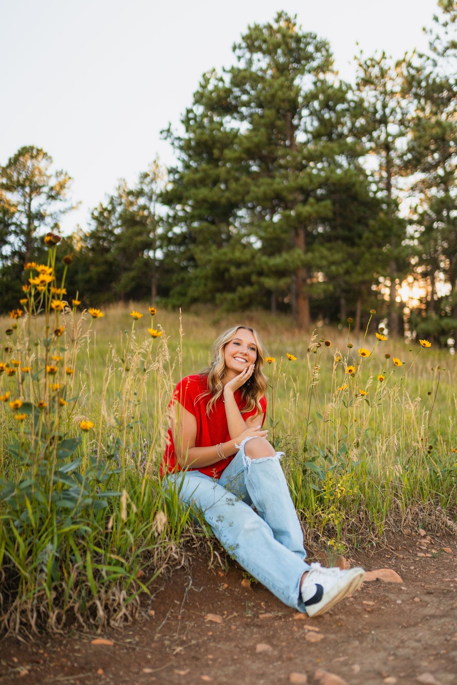 Person sitting in a field of yellow wildflowers at sunset wearing a red shirt and light jeans against a pine forest backdrop.