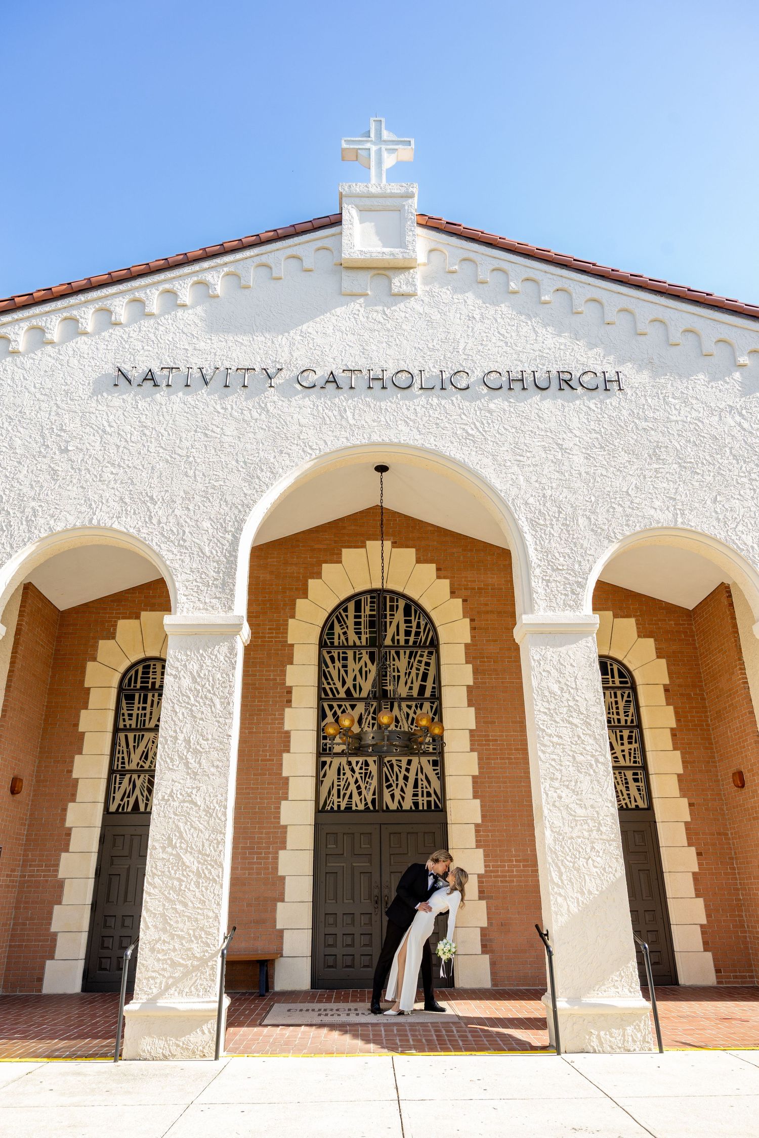 Couple sharing a kiss in front of Nativity Catholic Church under a bright blue sky.