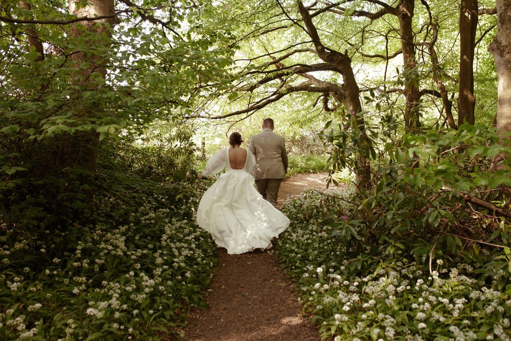 A couple walks hand in hand down a forest path surrounded by white wildflowers.