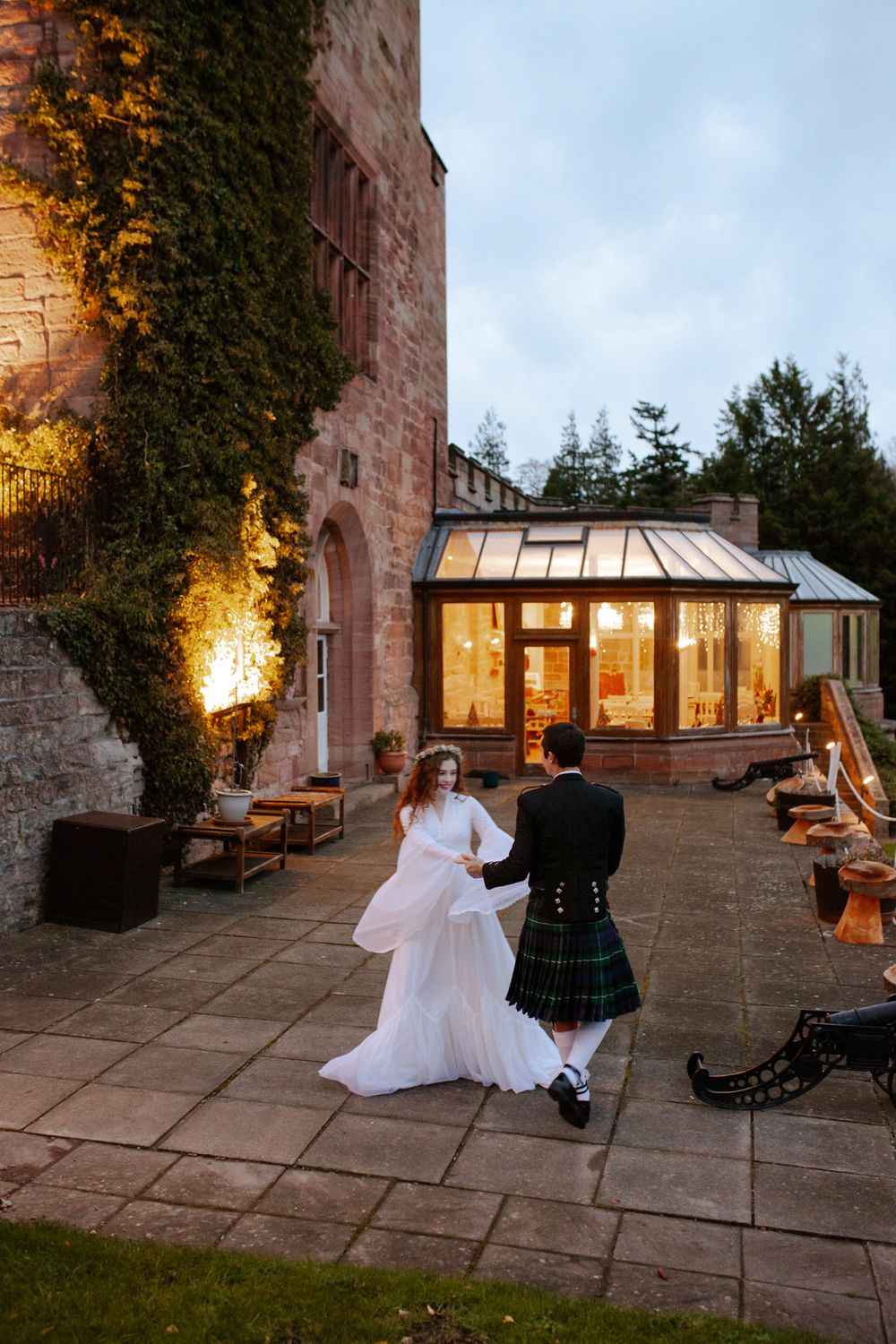 A couple dances on a castle terrace at dusk with the groom wearing a traditional Scottish kilt.