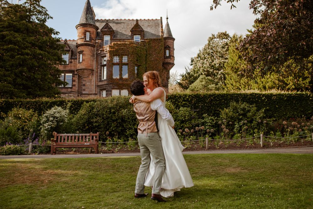 A couple dances together on a lush green lawn in front of a grand Victorian-style mansion during a wedding celebration in Edinburgh Zoo.
