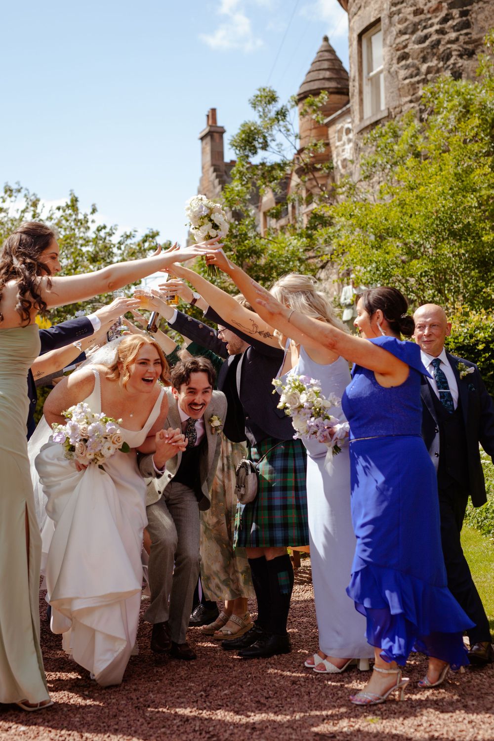 Wedding party celebrates outside historic stone castle with guests forming celebratory arch on sunny day.