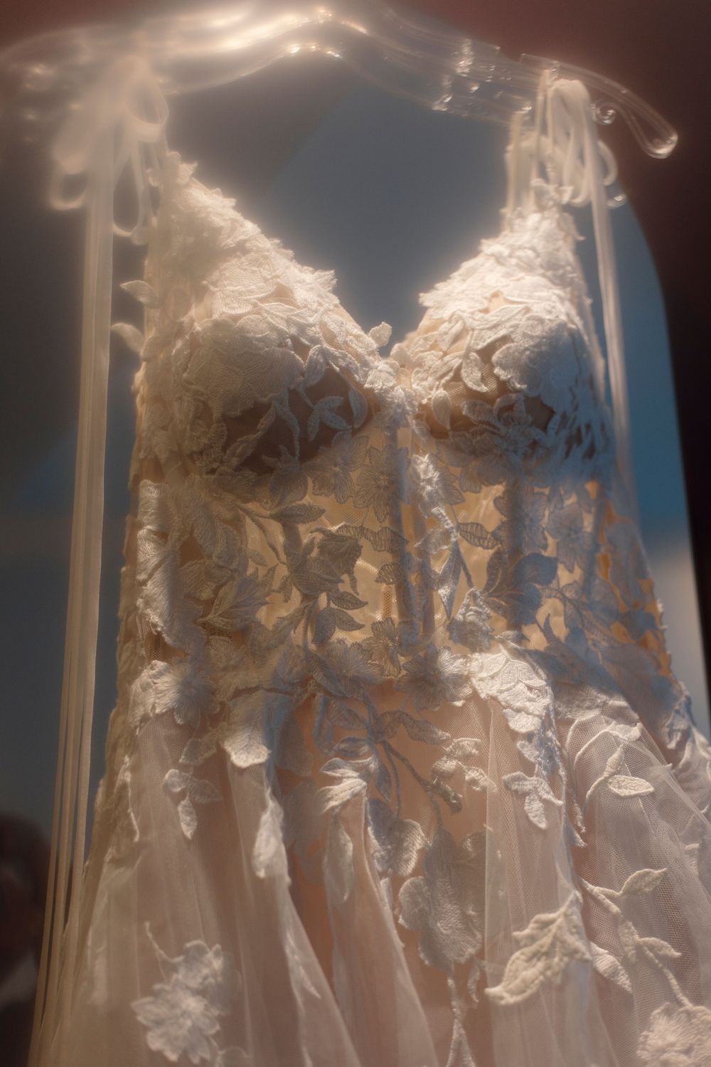 Ethereal white lace wedding dress with intricate floral appliques hanging on display in soft romantic lighting.
