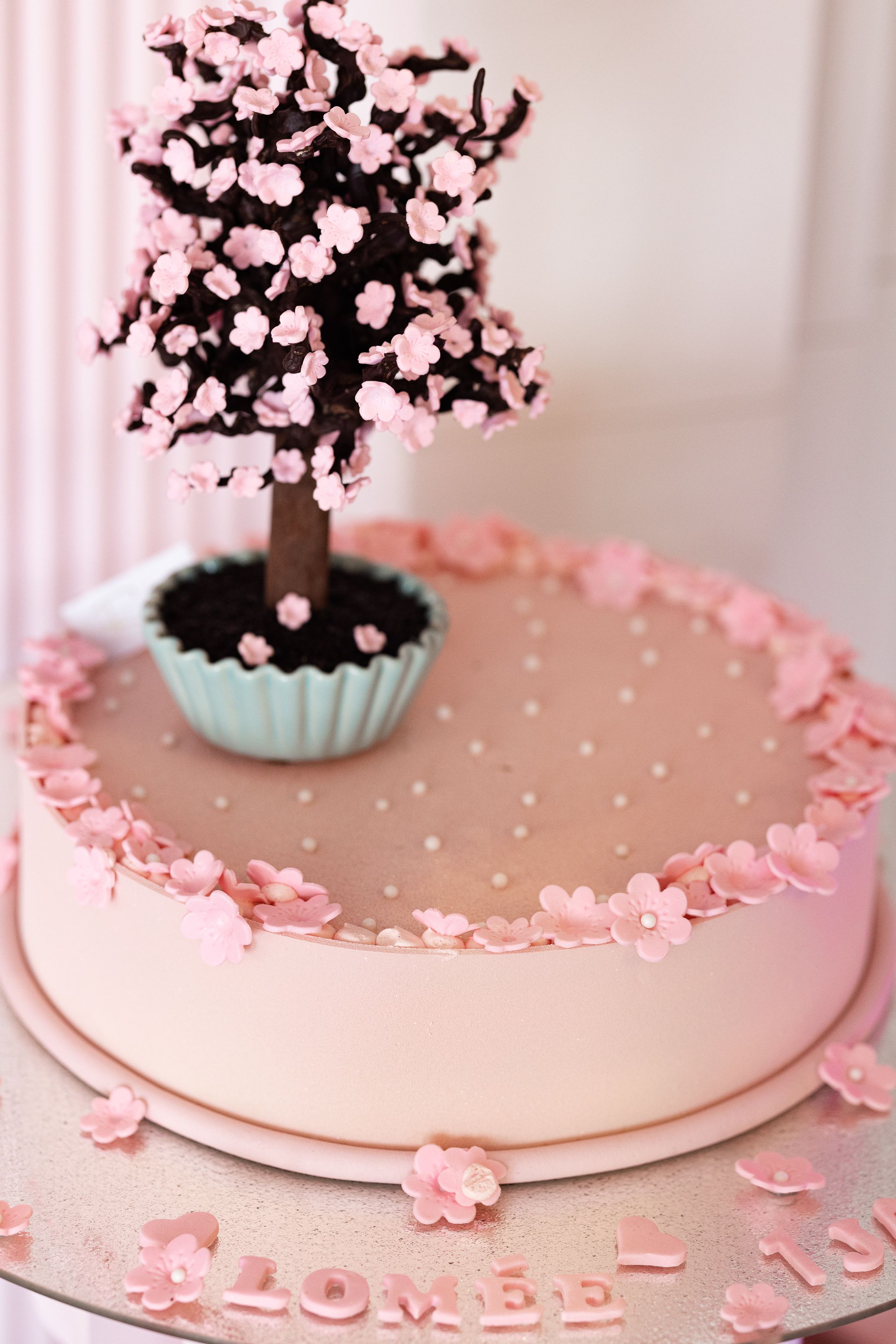Pink floral cake decorated with cherry blossoms and a turquoise vase on a white pedestal stand.