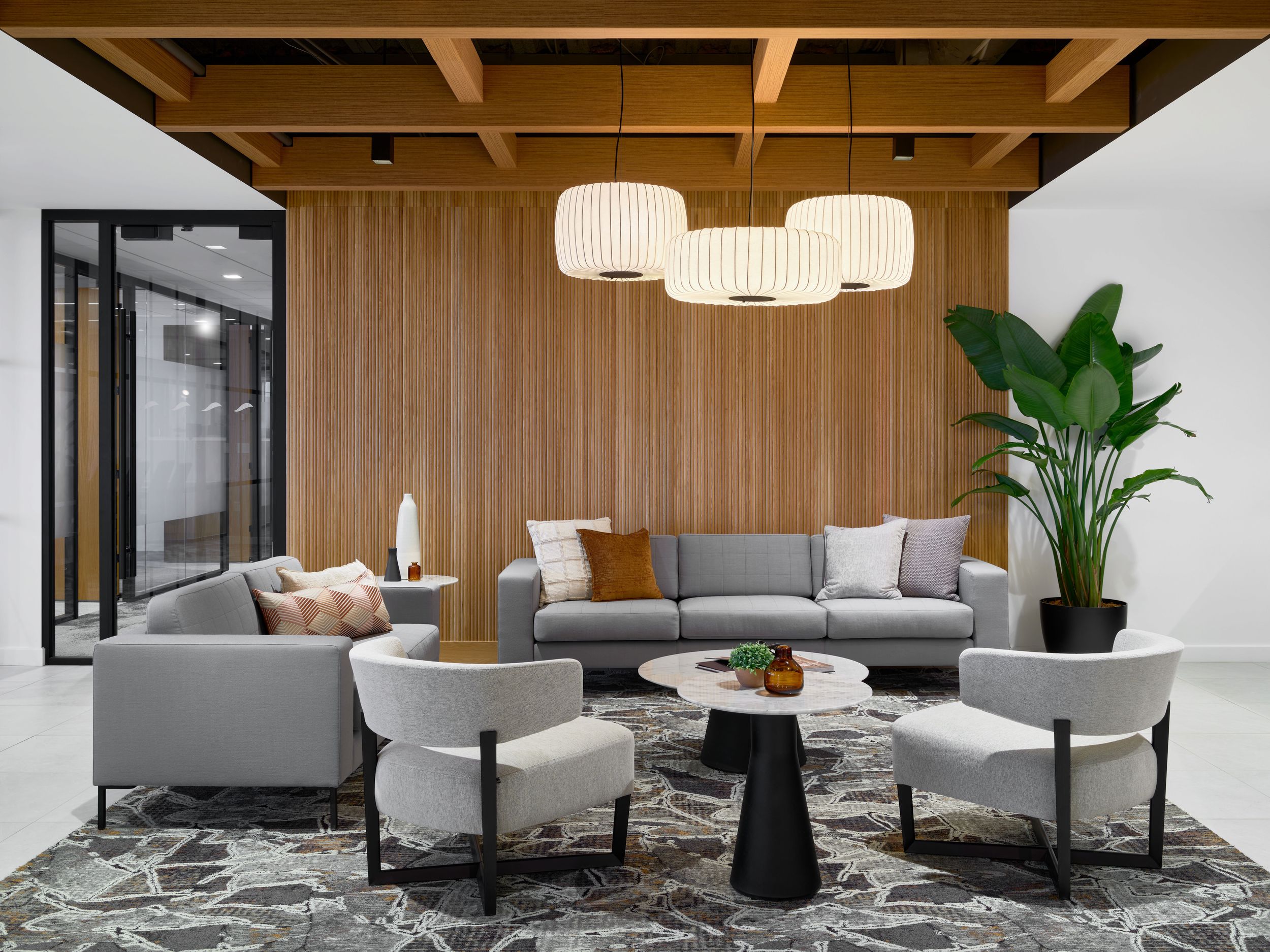Contemporary office lounge area features wood paneling, modern lighting, and gray sectional seating with indoor plants.