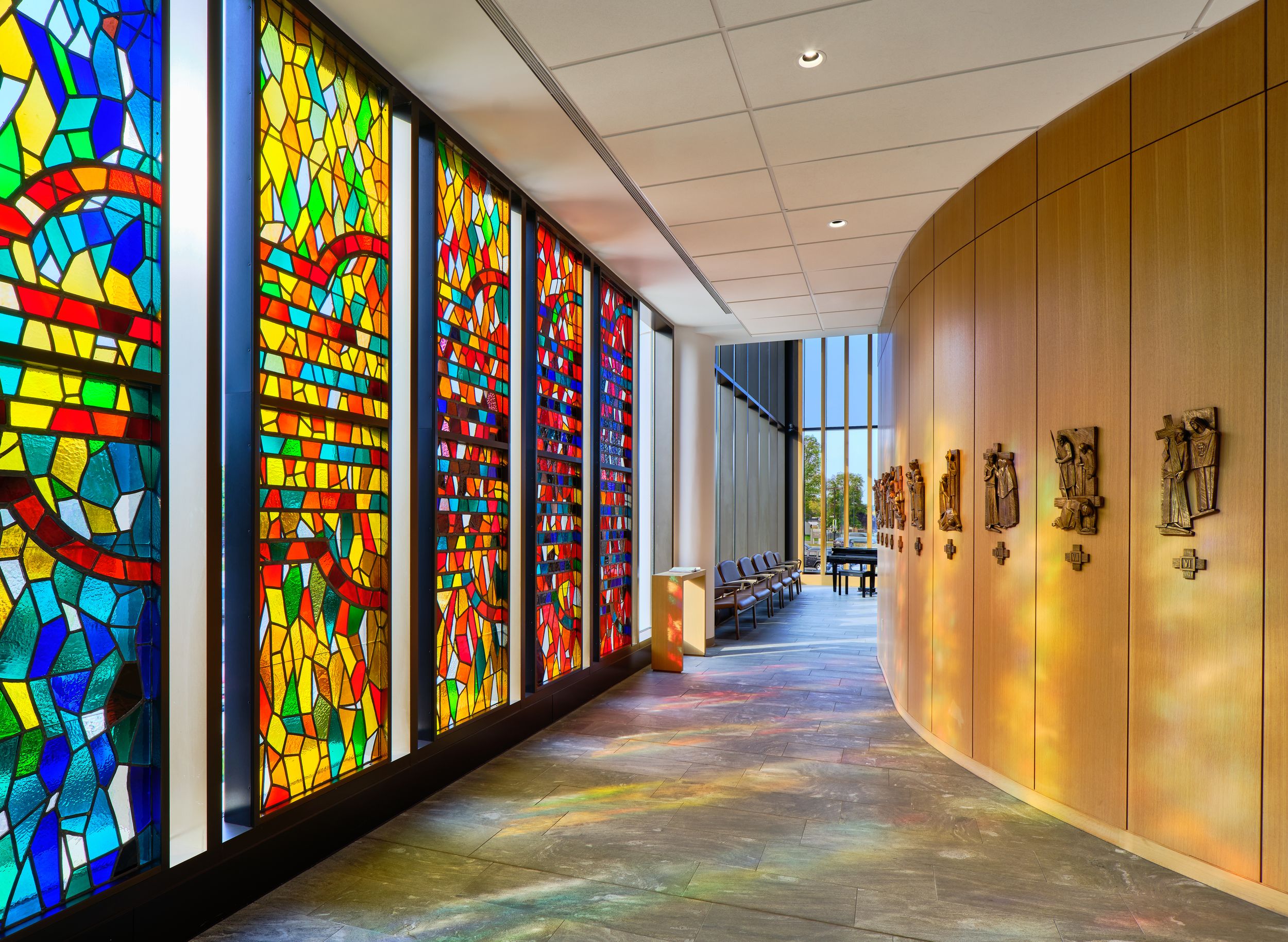 A curved hallway features colorful stained glass windows alongside wooden paneled walls in a modern architectural setting.