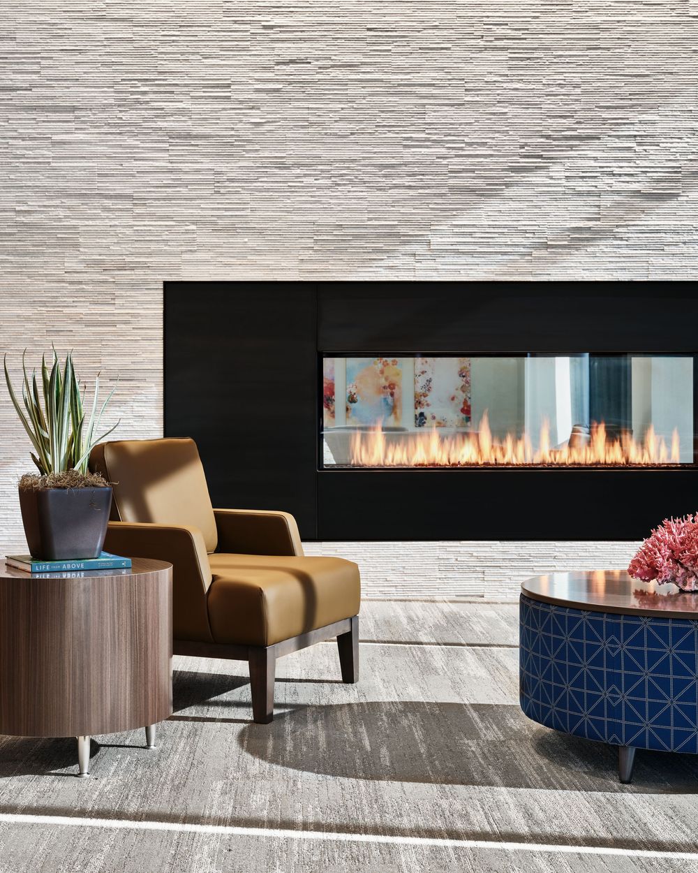 Modern living room with luxurious fireplace, tan armchair, round side table, potted plant, and geometric blue ottoman.