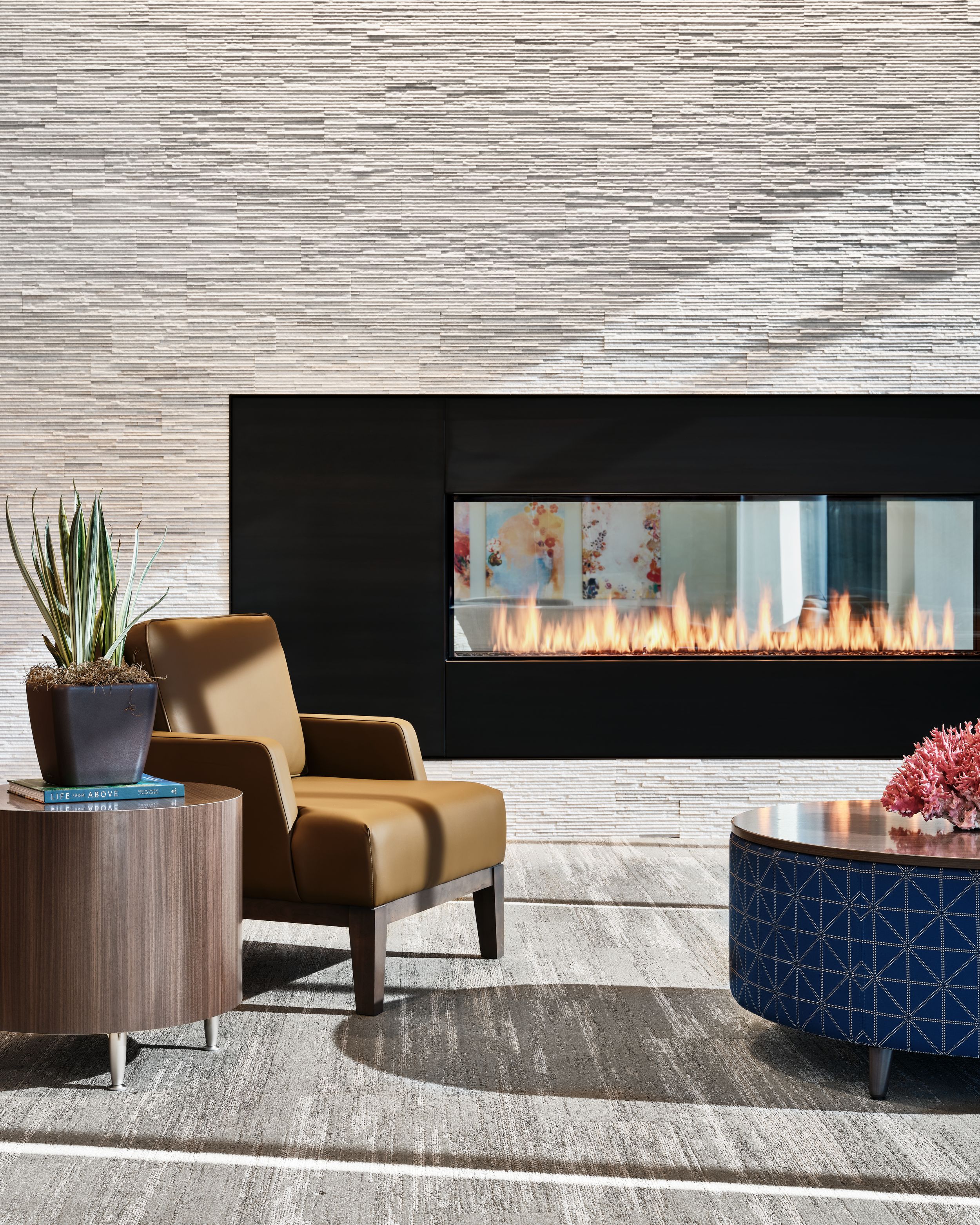 Modern living room with luxurious fireplace, tan armchair, round side table, potted plant, and geometric blue ottoman.