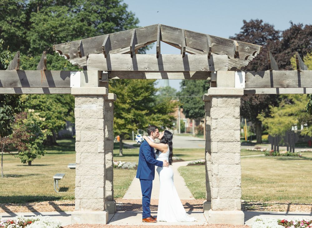 A couple stands beneath an elegant stone pergola in a serene garden setting during their wedding ceremony.