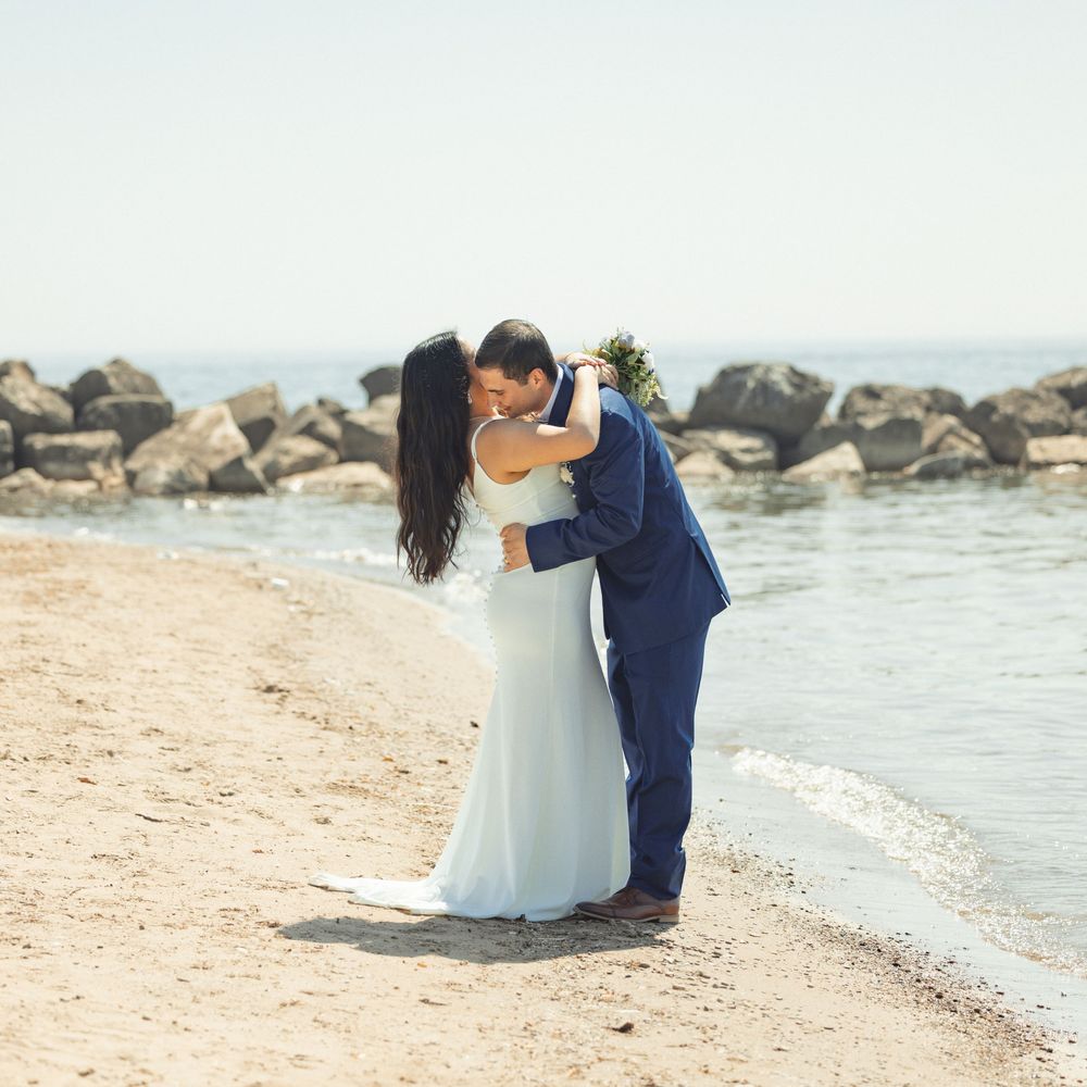 A couple in wedding attire embrace on a rocky beach with ocean waves in the background.