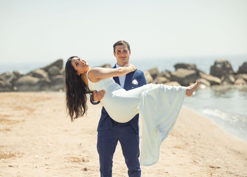 Two people in matching blue outfits playfully run and dance together along a sunny beach with ocean waves in the background.