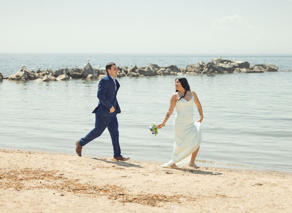 A couple in wedding attire runs playfully along a sandy beach with rocky coastline in the background.