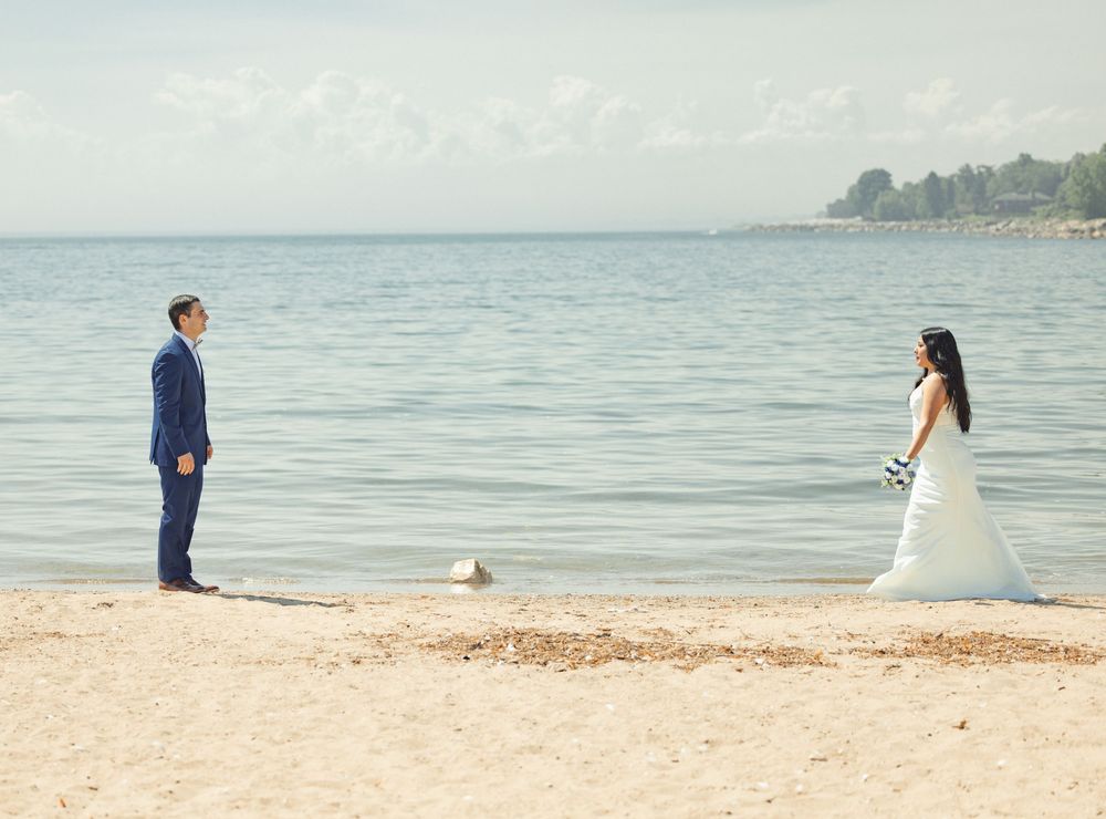 A couple stands apart on a tranquil beach with calm waters and soft sand during an elegant beachside moment.