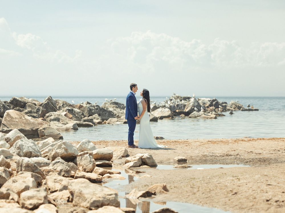 A couple standing on rocky coastal shoreline with calm ocean waters and blue sky in the background.